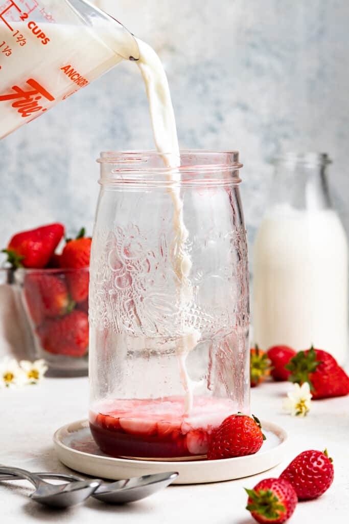 Pouring milk into a mason jar with strawberry syrup. Fresh strawberries and milk are in the background.