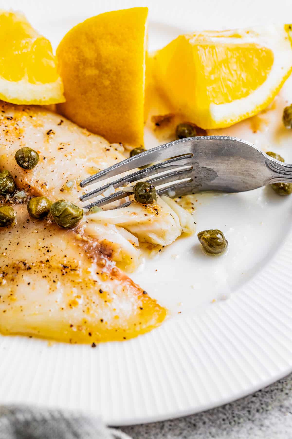 Air fryer tilapia on a plate with lemon caper sauce and fresh lemons and a fork taking a bite out of it.