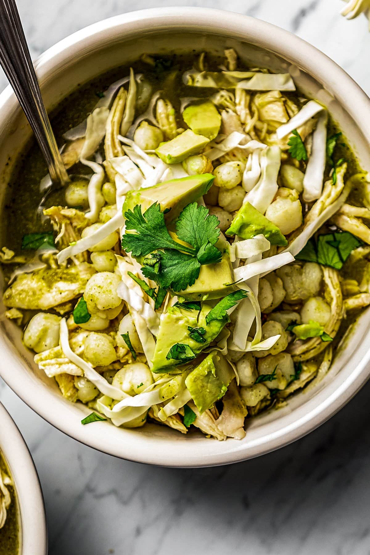 close up of chicken pozole verde served in a bowl and garnished with shredded cabbage, avocado slices, and fresh cilantro.