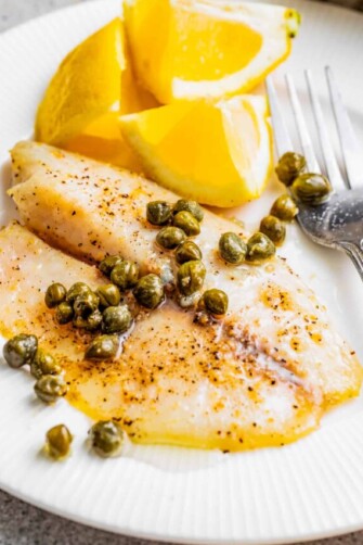 Air fryer tilapia on a plate with lemon caper sauce, fresh lemons, and a fork.