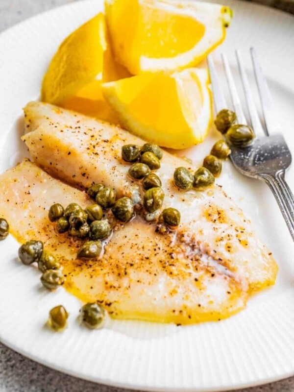 Air fryer tilapia on a plate with lemon caper sauce, fresh lemons, and a fork.