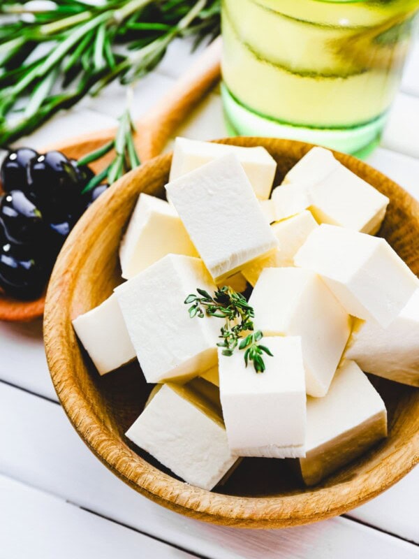 Feta cheese cubes in a brown wooden bowl, with a rosemary sprig, olive oil, and olives set behind the bowl.