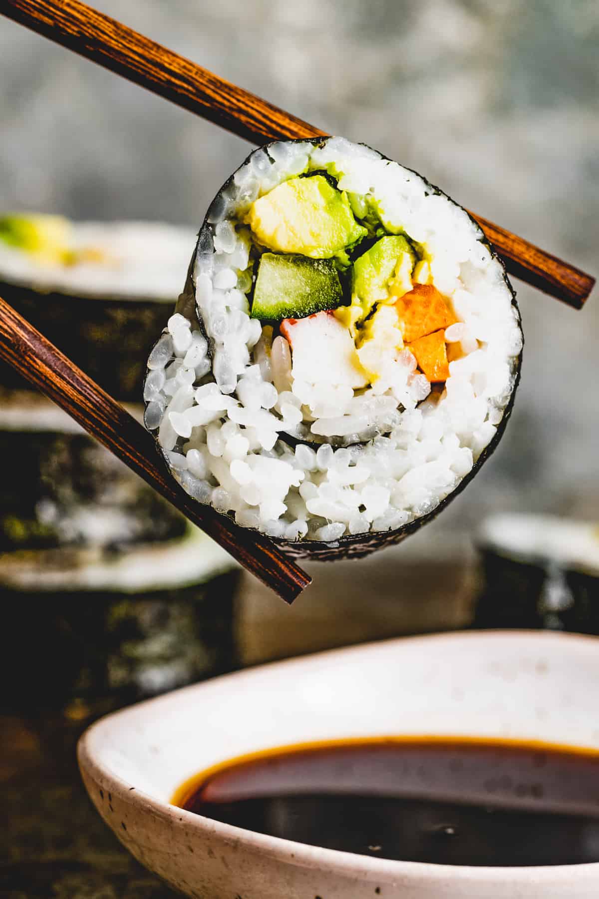 Chopsticks holding a slice of maki sushi over a bowl of soy sauce.