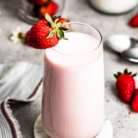 Strawberry milk in a glass with a strawberry on the edge of the glass, and fresh strawberries arranged around the glass of milk.