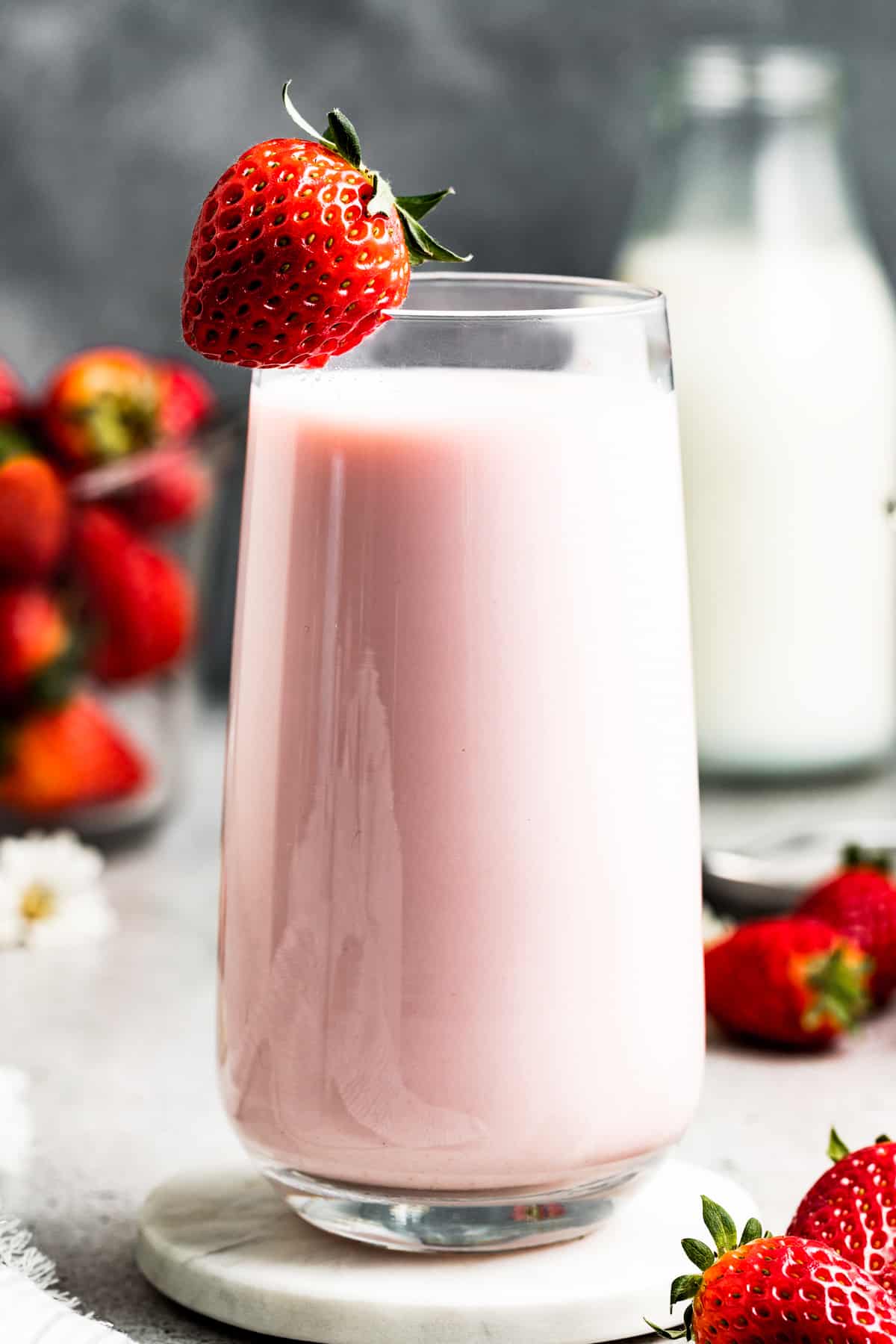Strawberry milk in a glass with a strawberry on the edge of the glass. Fresh strawberries and milk are placed behind the milk glass.