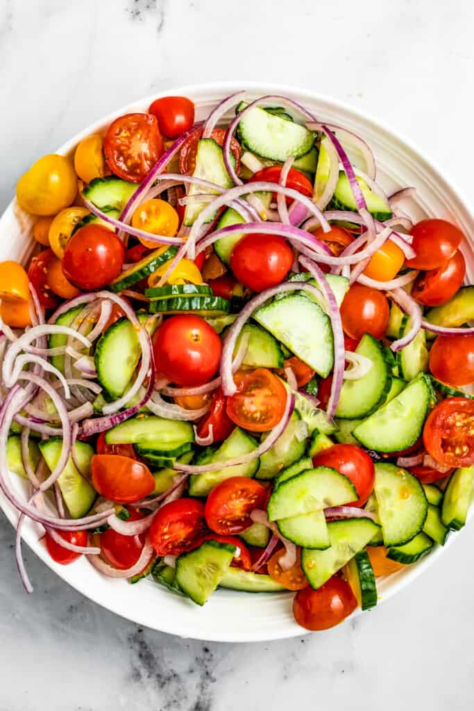 A round salad bowl filled with sliced cucumbers, cherry tomatoes, and sliced red onions.