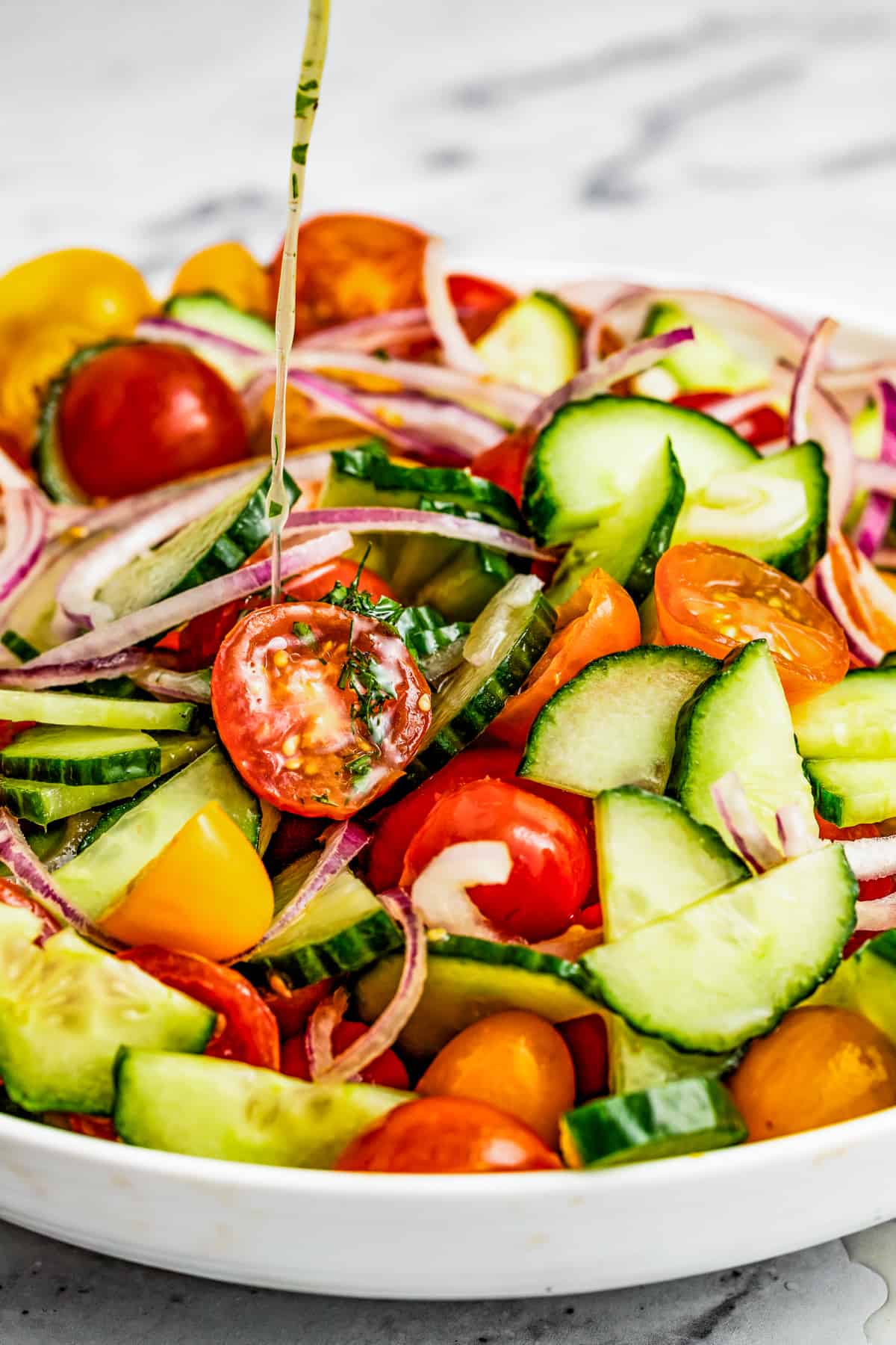 A side shot of a plate with cucumber tomato salad and a stream of dressing poured over it.