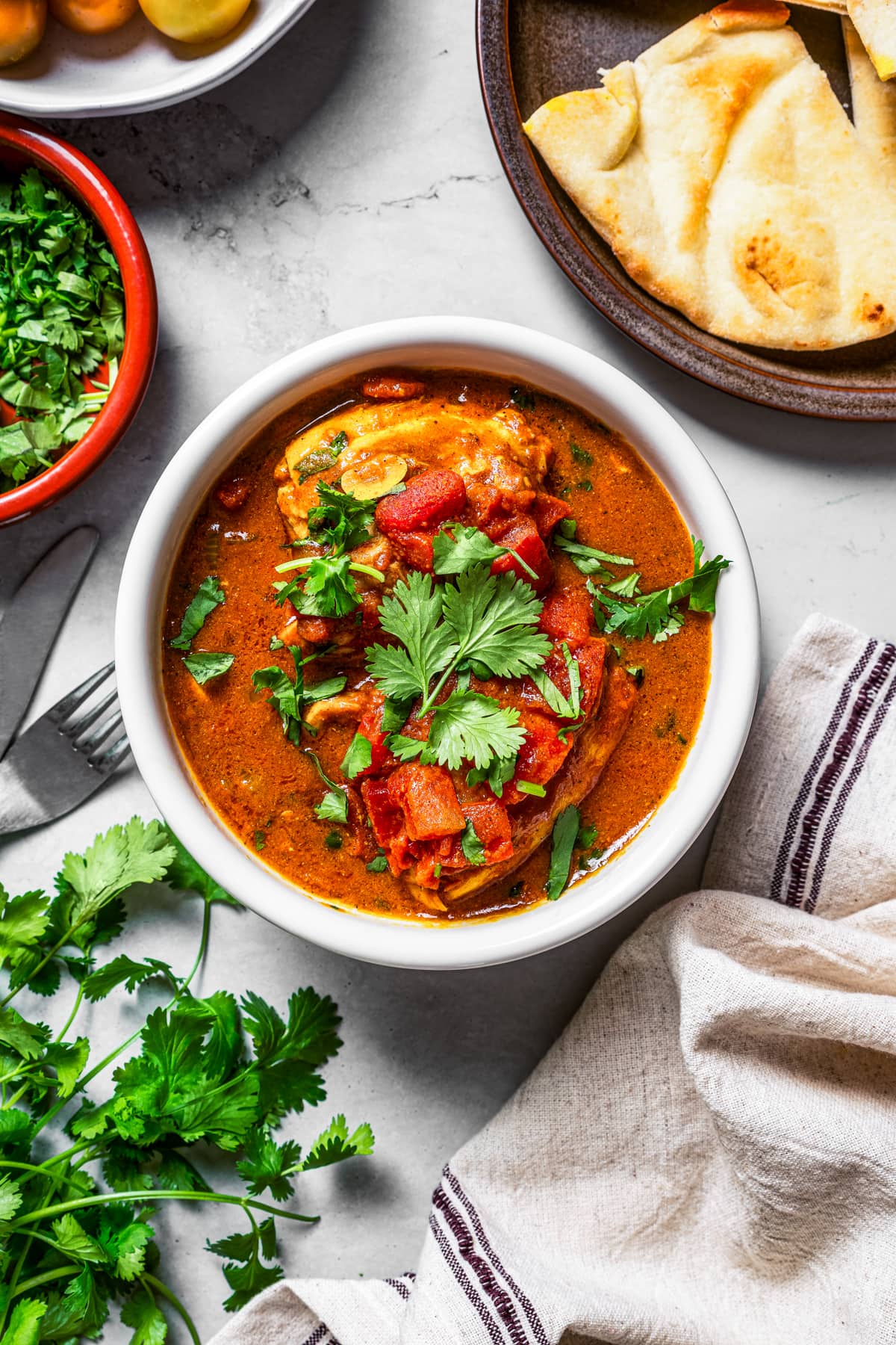 Overhead of a bowl of chicken rogan josh near fresh cilantro and naan.