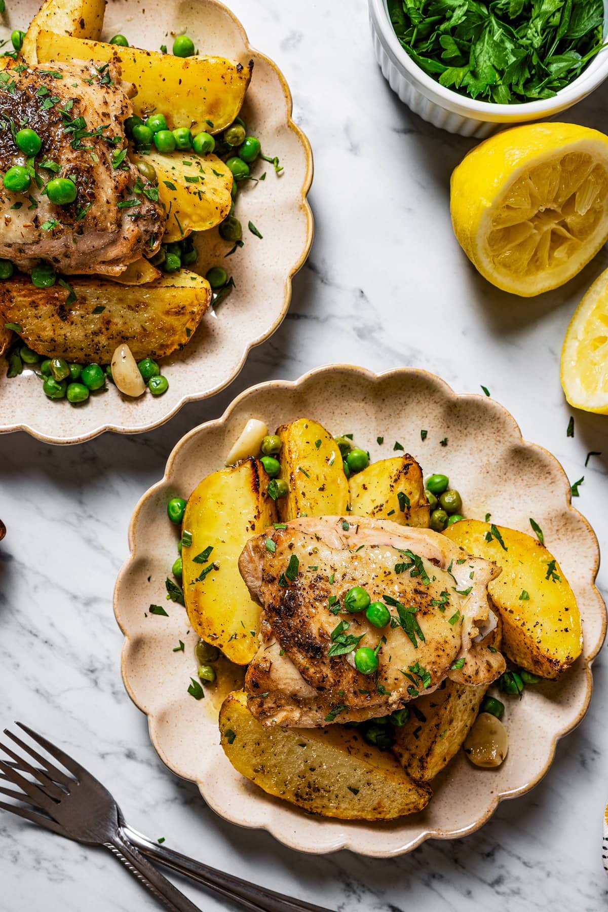 Overhead shot of two plates of chicken Vesuvio near a squeezed fresh lemon and fresh parsley.