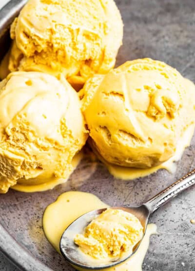 Three scoops of mango ice cream in a pan with a spoon that has mango ice cream in it.