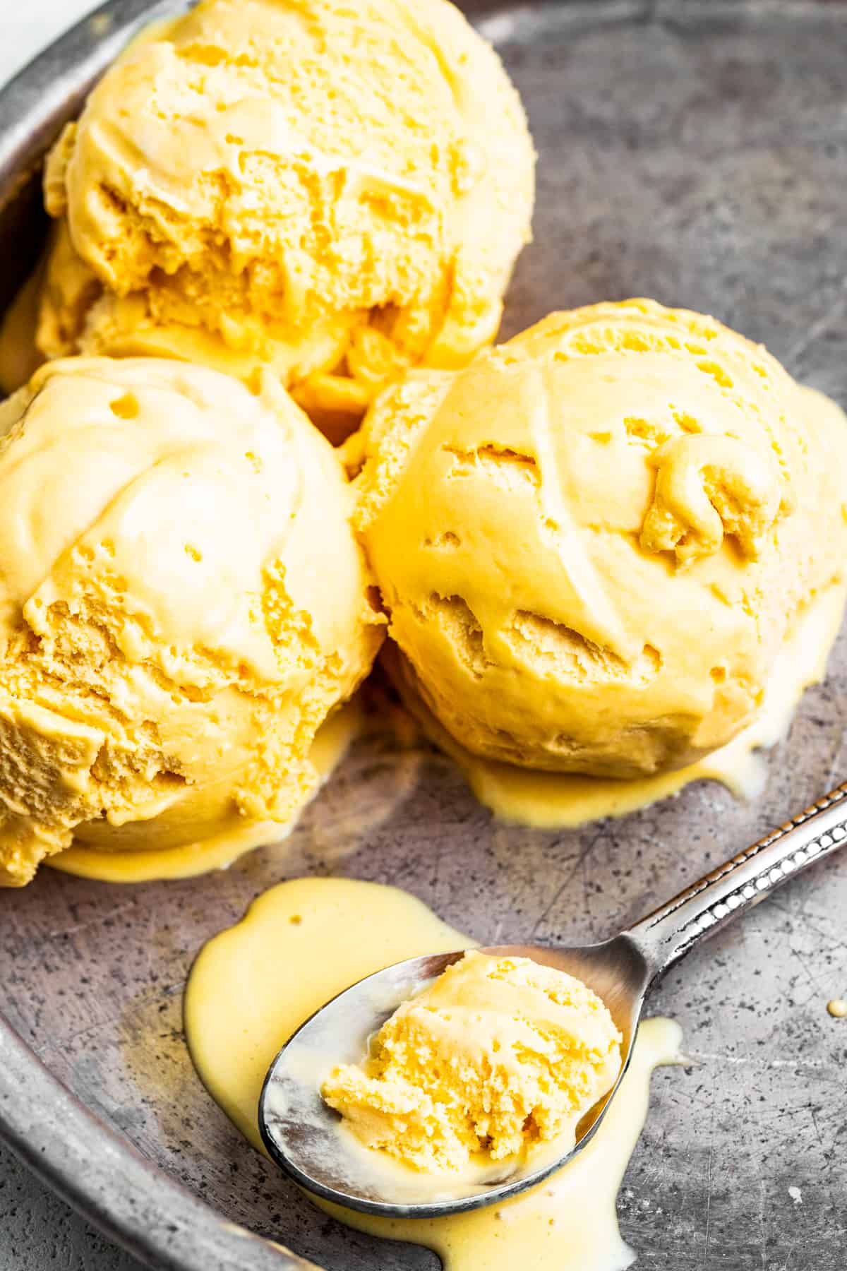 Three scoops of mango ice cream in a pan with a spoon that has mango ice cream in it.