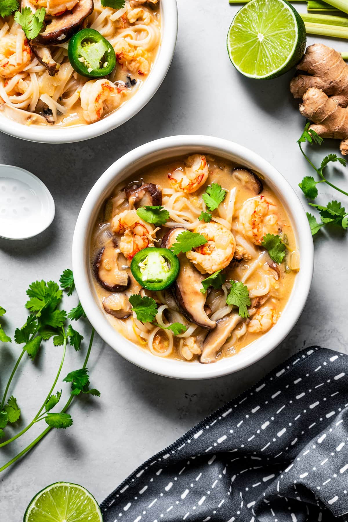 Overhead image of tom yum noodle soup served in two bowls surrounded by lime, cilantro, ginger, and lemongrass.