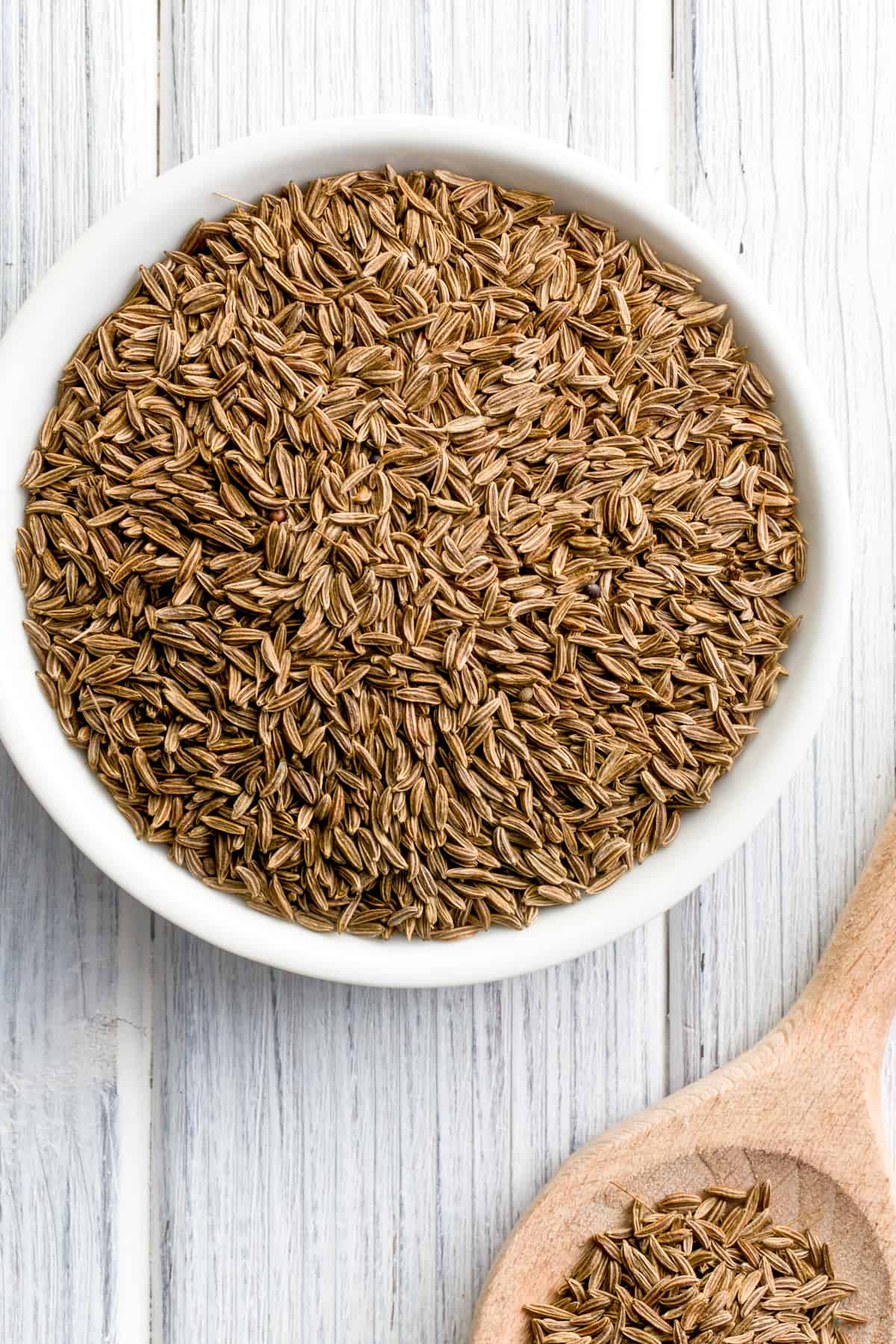 Cumin seeds in a bowl, with a wooden spoon also holding cumin seeds right next to the bowl.