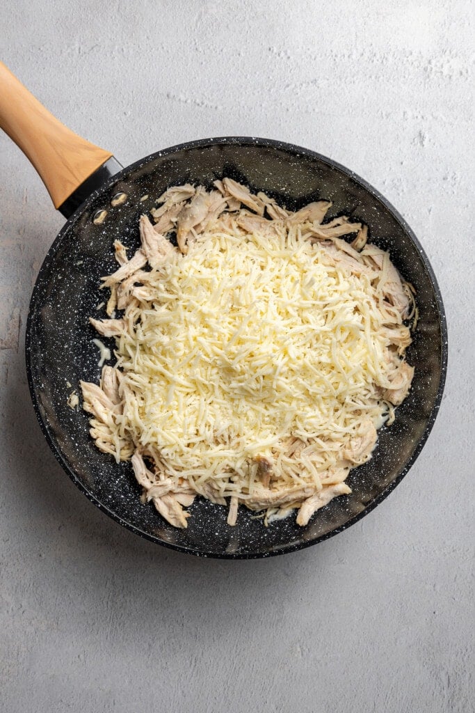 Adding cheese to shredded chicken in a pan.