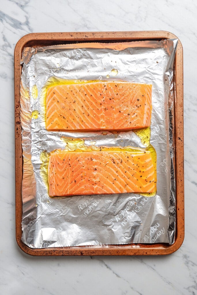Coating salmon with olive oil and seasoning it with salt and pepper on a sheet pan lined with aluminum foil.