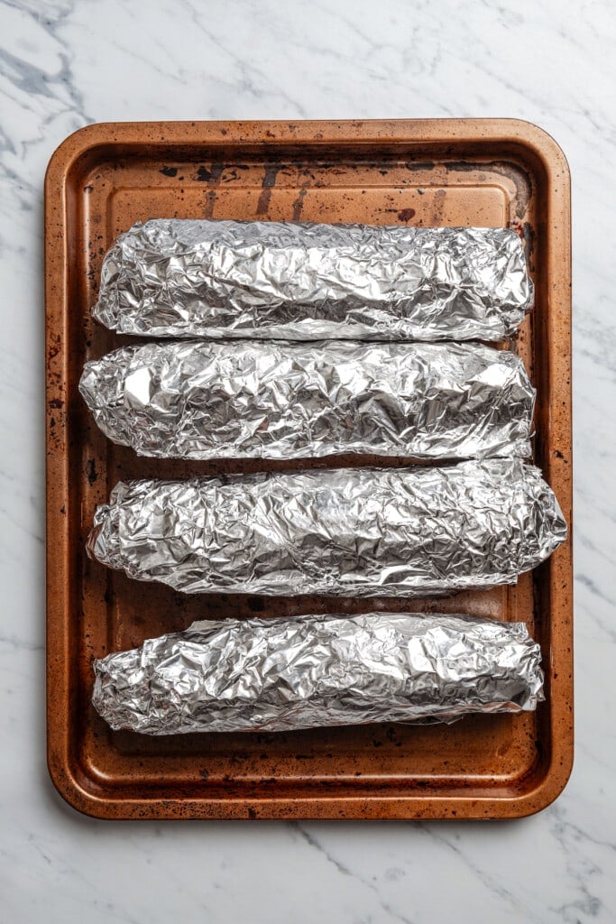 Four ears of corn wrapped in foil on a baking sheet.
