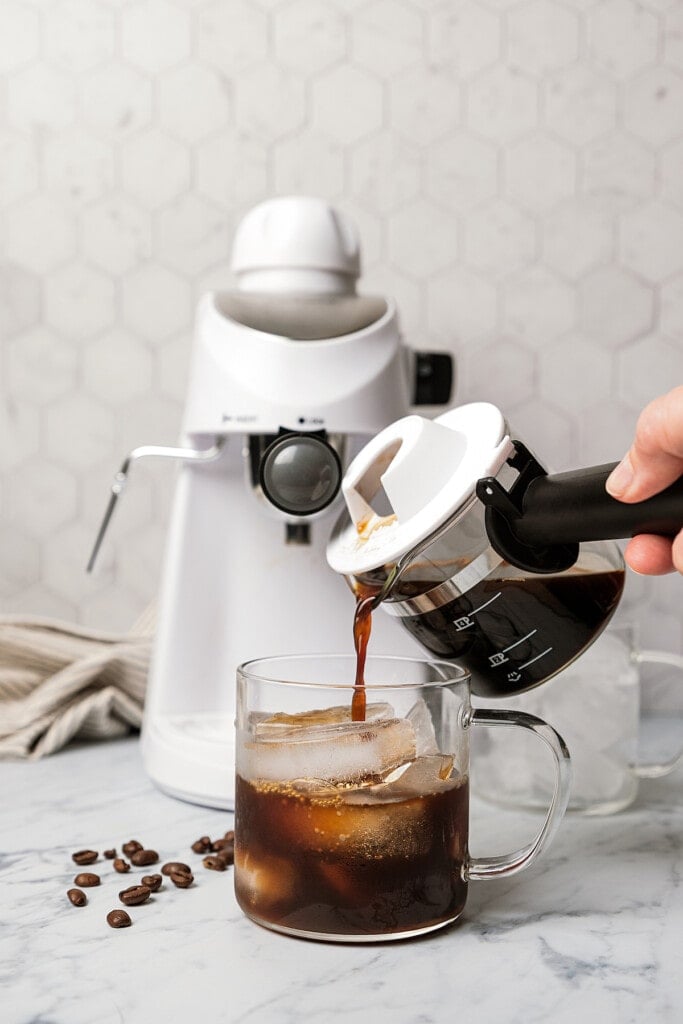 Brewed espresso is poured over a glass of ice, with the espresso machine in the background.