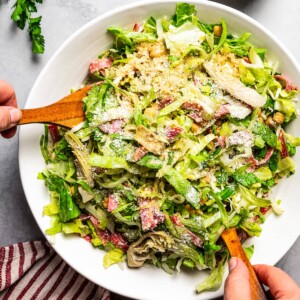 Hands using two wooden salad scoops to toss a La Scala chopped salad in a large serving bowl.