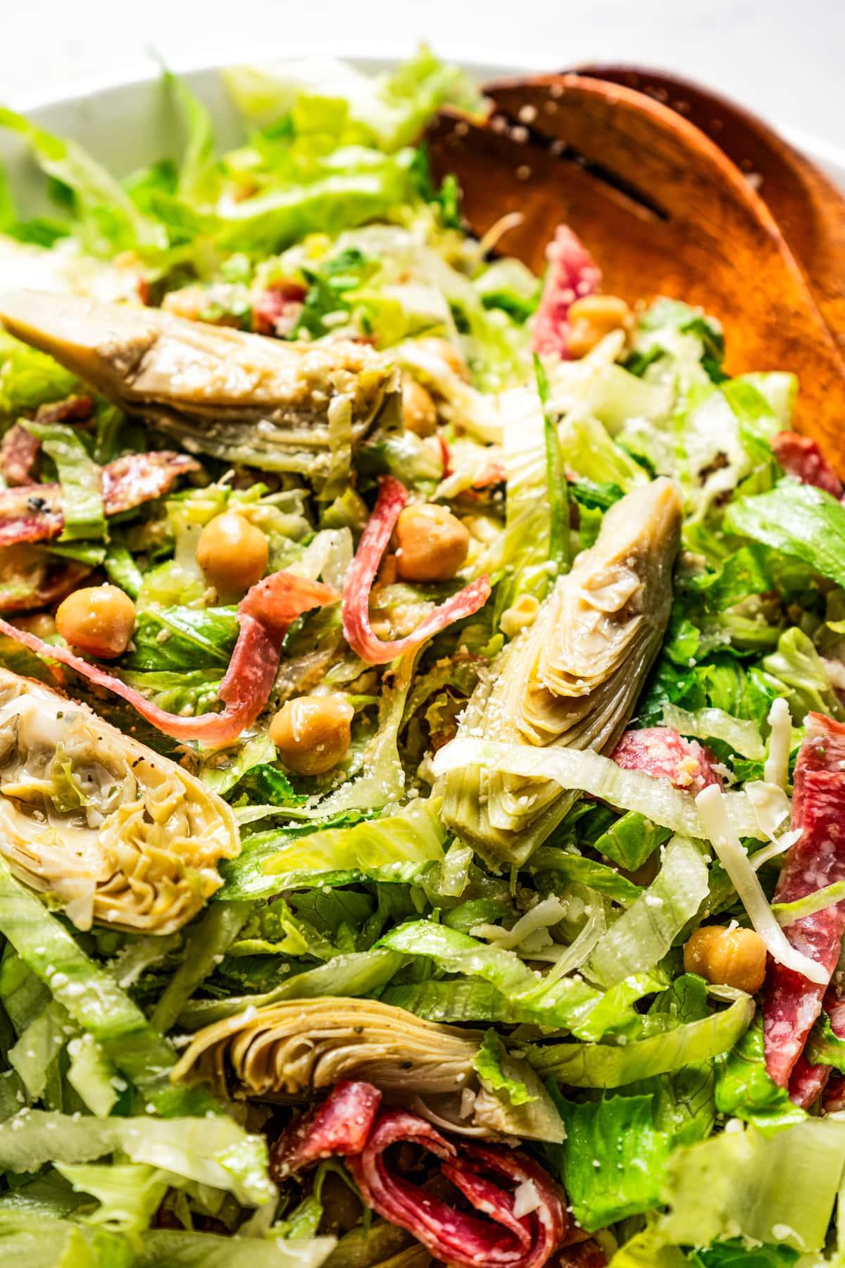Close-up photo of a La Scala chopped salad in a large bowl with sliced artichokes, salami, and chickpeas, next to two wooden serving spoons.