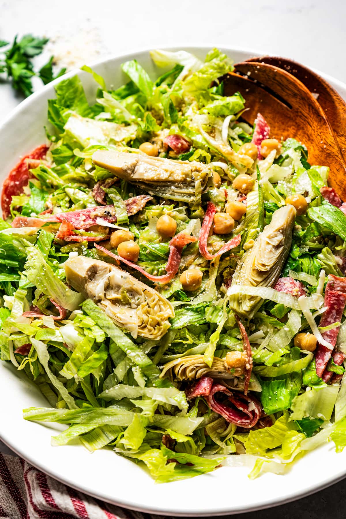 Image of La Scala chopped salad in a large bowl with sliced artichokes, salami, and chickpeas, next to two wooden serving spoons.