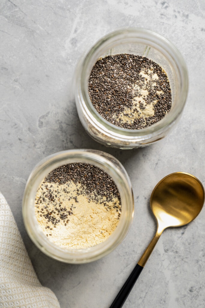 Overhead view of chia seeds and protein powder added to oats in two mason jars next to a gold spoon.