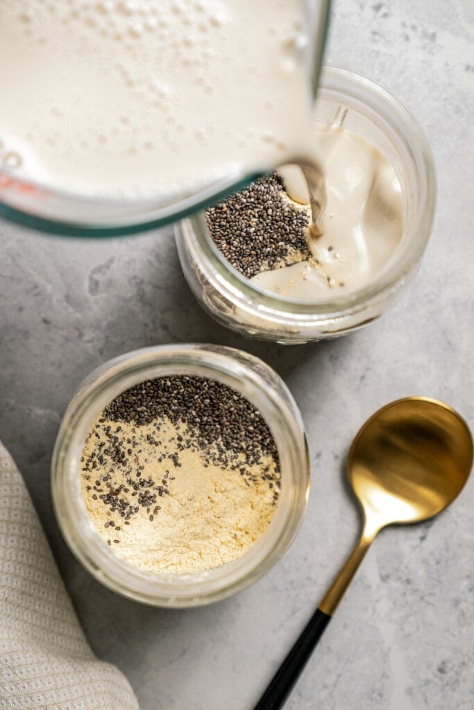 Milk is poured into mason jars filled with oats, chia seeds, and protein powder.