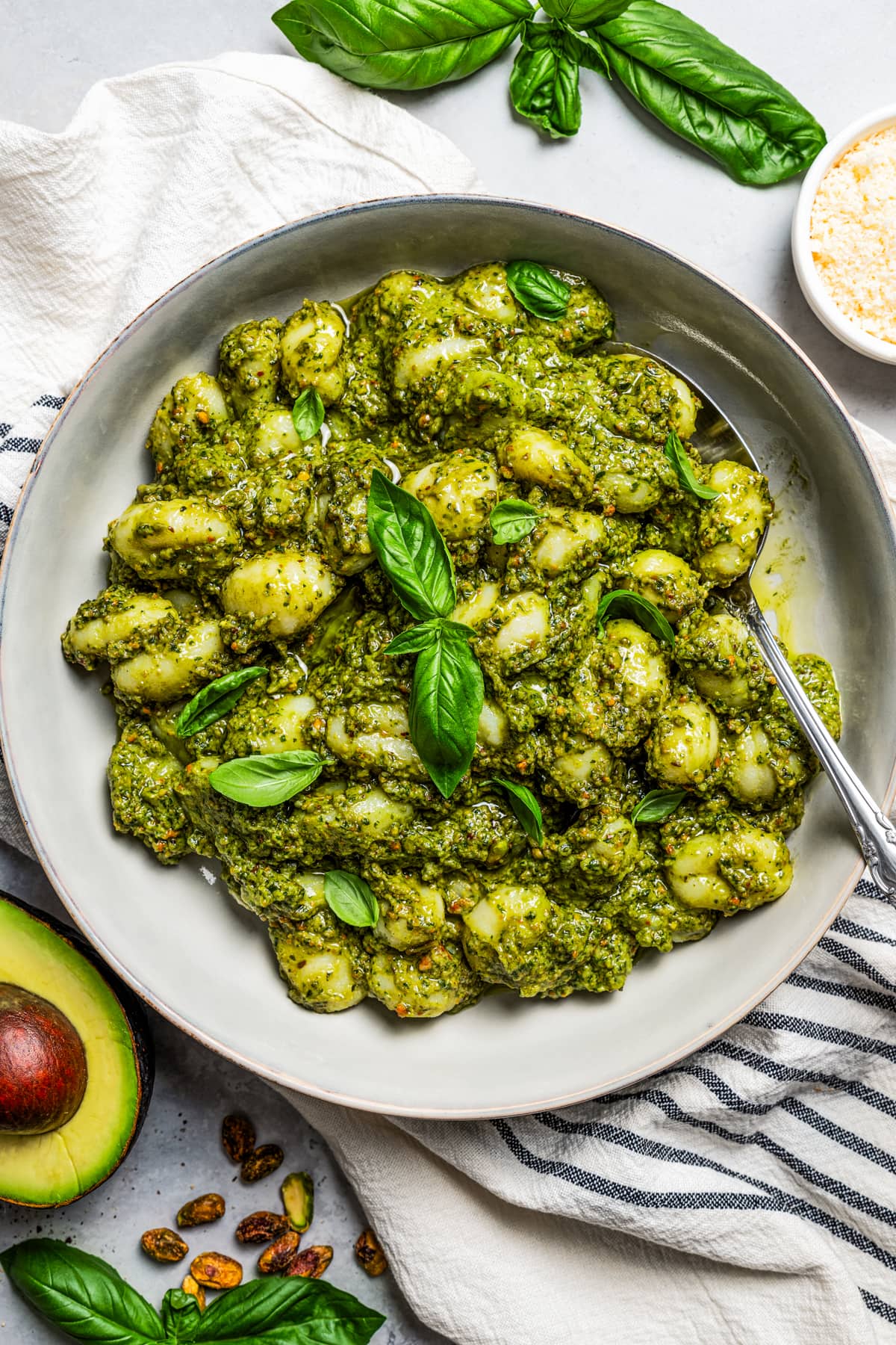 Overhead image of avocado pesto pasta served on a plate, with a spoon resting on the plate, and a halved avocado placed next to the plate.