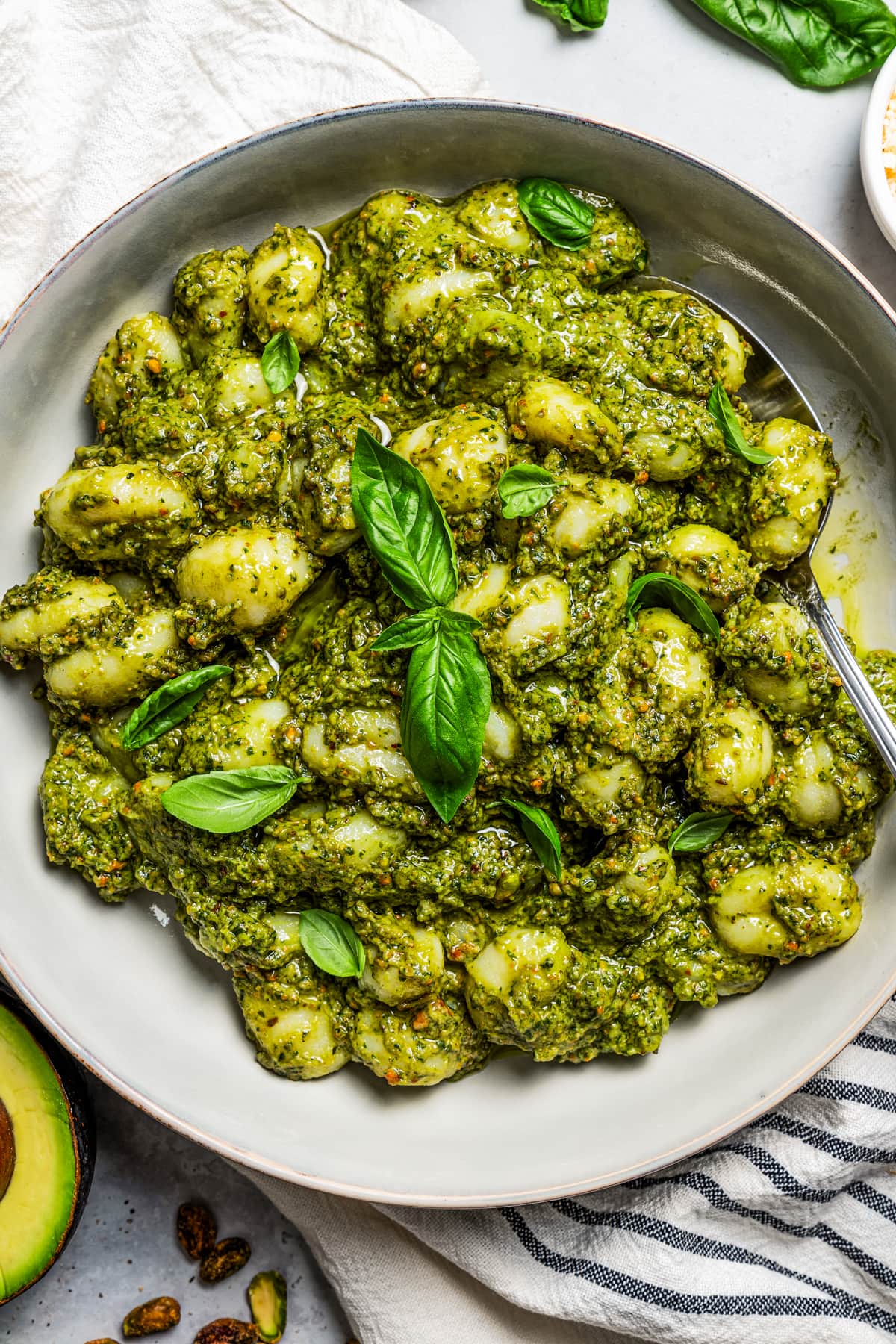 Overhead image of avocado pesto pasta served on a plate, with a spoon resting on the plate, and a halved avocado placed next to the plate.