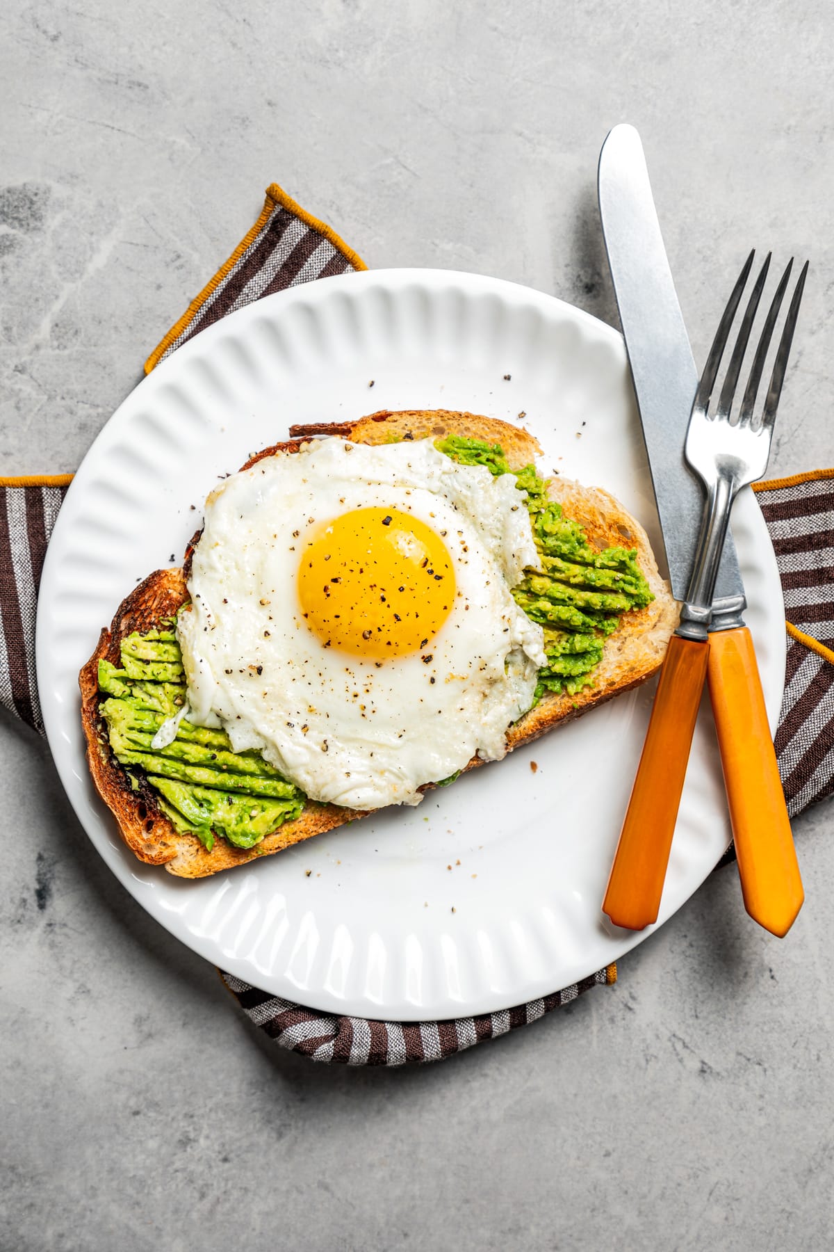 Avocado toast with an egg on top on a plate with a fork and knife.