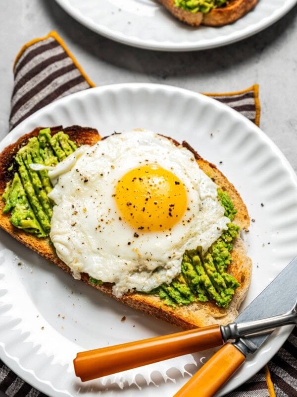 Avocado toast with an egg on top on a plate with a fork and knife. A cup of iced coffee sits nearby.