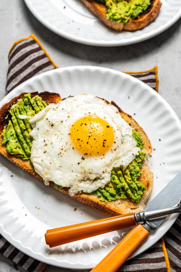 Avocado toast with an egg on top on a plate with a fork and knife. A cup of iced coffee sits nearby.
