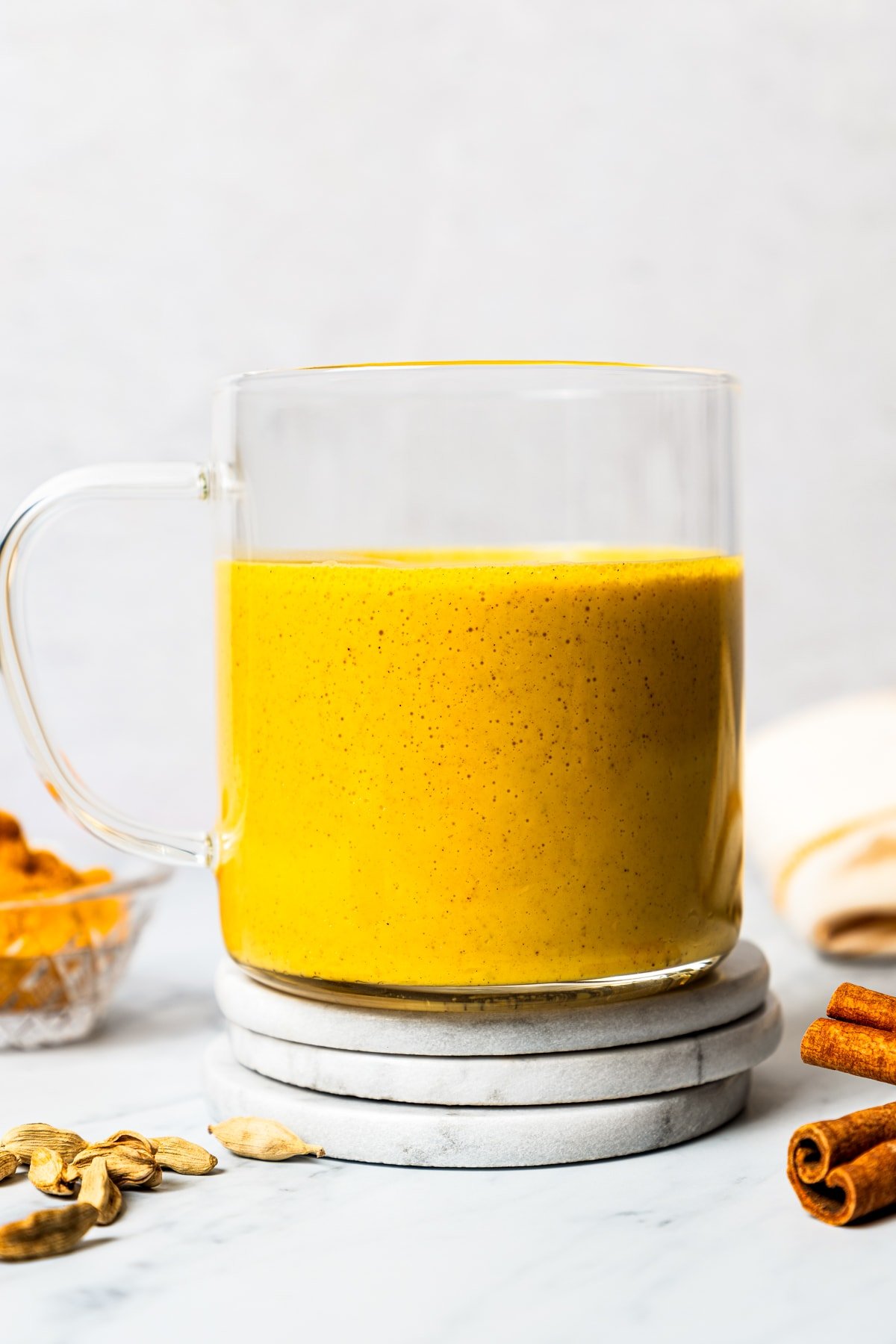 A glass mug filled with turmeric latte near a bowl of turmeric and other whole spices.
