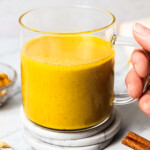 A hand holding a glass mug filled with turmeric latte near a bowl of turmeric and cinnamon sticks.