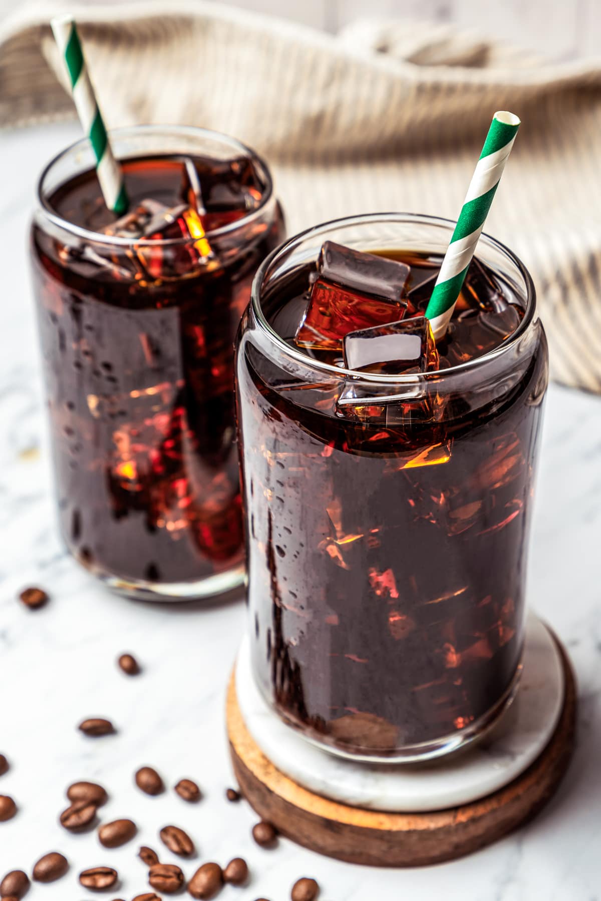 Iced Americano drinks on a wooden coaster with another glass of coffee set behind it and espresso grounds scattered around the drinking glasses.