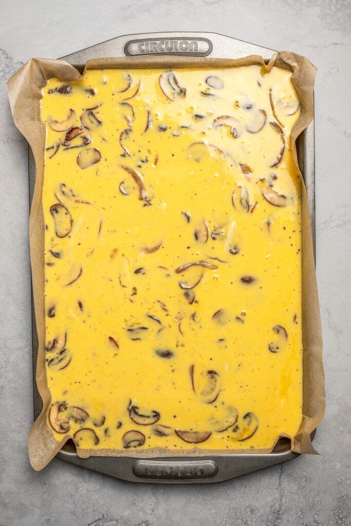 Pouring scrambled eggs mixed with sautéed mushrooms, onions, half and half, and cheeses into a sheet pan to bake.