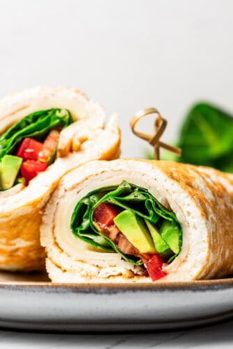 Closeup of a stuffed egg white wrap rolled up with cheese, turkey, avocado, salad, and tomatoes, and served on a plate.