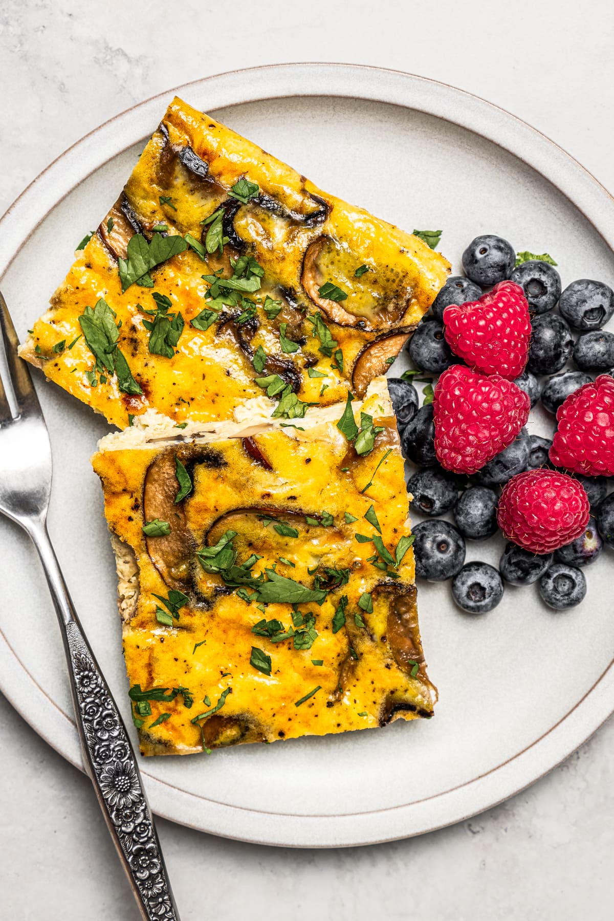 Slices of sheet pan eggs served on a plate with berries. Fresh berries are scattered around and another plate of sheet pan eggs is nearby.