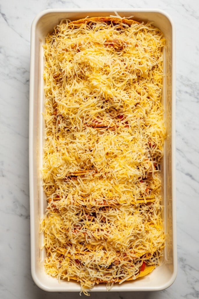 Sprinkling shredded shredded Mexican cheese over tacos in a baking dish.