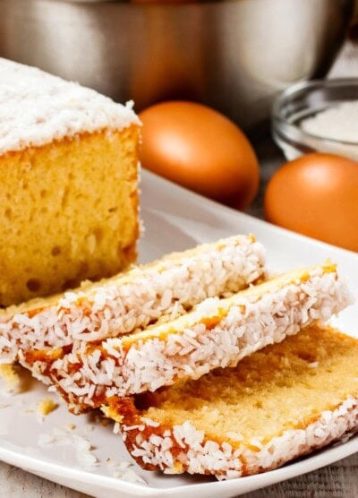 Banana pound cake topped with coconut flakes, arranged on a white platter, and partially sliced.