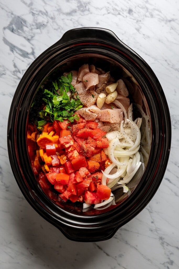 Loading a crockpot with chicken, veggies, herbs, seasonings, and chicken broth.