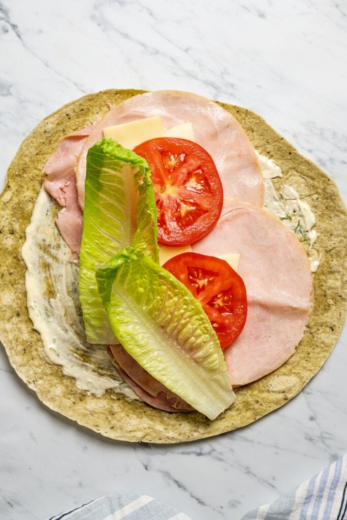 Adding lettuce to a wrap spread with dijon-dill mayo and filled with meats, cheeses, and tomato.