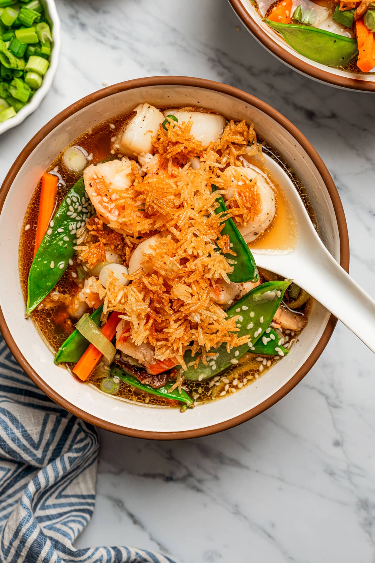 Overhead image of a soup bowl filled with broth, scallops, shrimp, and veggies, and topped with fried rice.