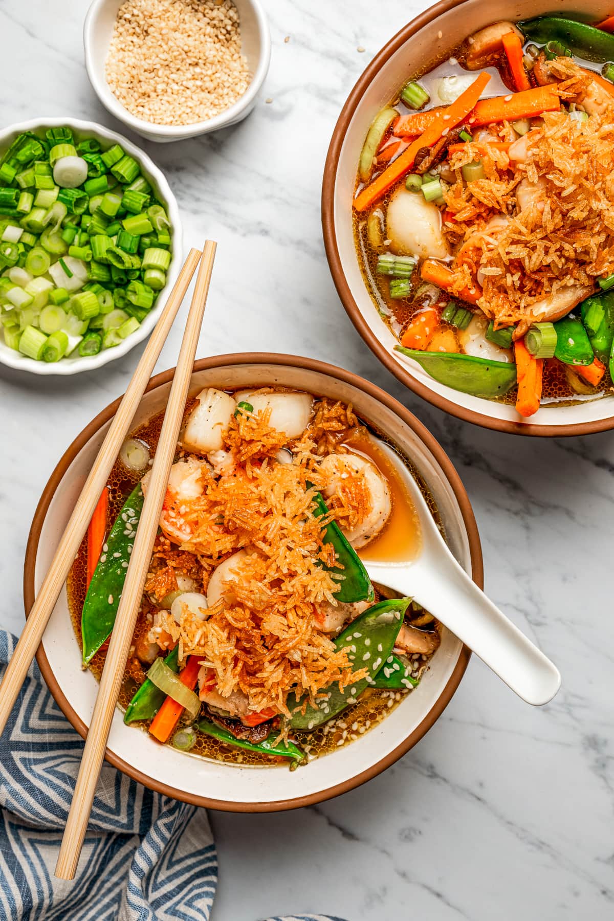 Overhead image of two soup bowls with broth, scallops, shrimp, and veggies, topped with fried rice, with chopsticks resting on one of the bowls.