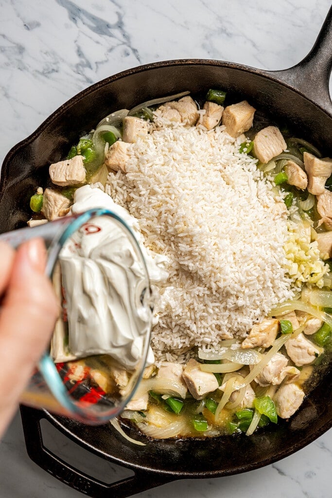 Adding garlic, rice, sour cream, and water to sauteed chicken and veggies in a skillet.