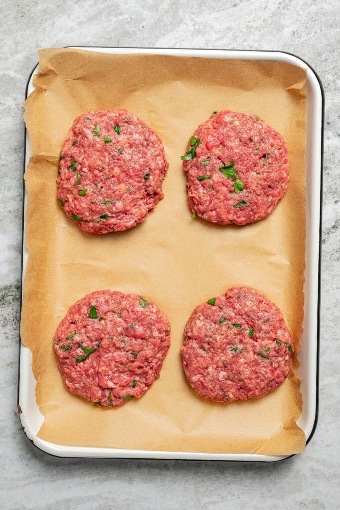 Seasoned beef patties on a baking sheet with parchment paper.