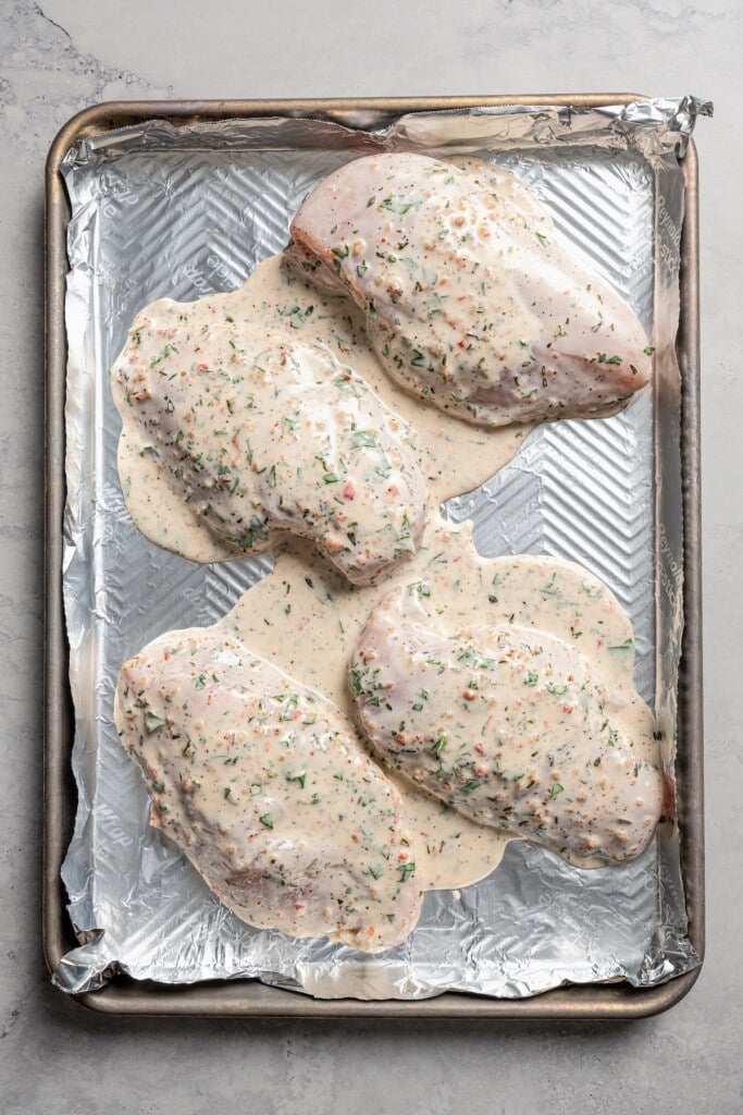 Pouring Italian dressing marinade over chicken breasts on a baking sheet lines with aluminum foil.