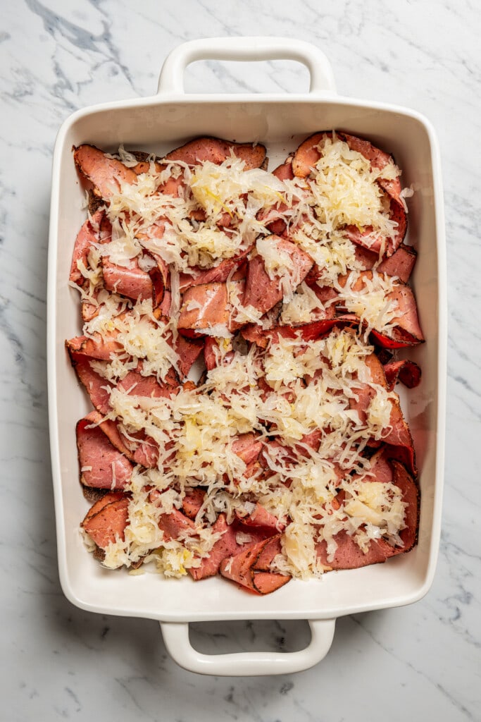 Overhead image of a baking dish with shredded Swiss cheese layered over pastrami.