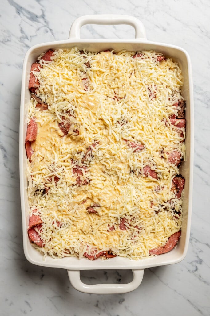Overhead image of a baking dish with shredded Swiss cheese and custard layered over pastrami.