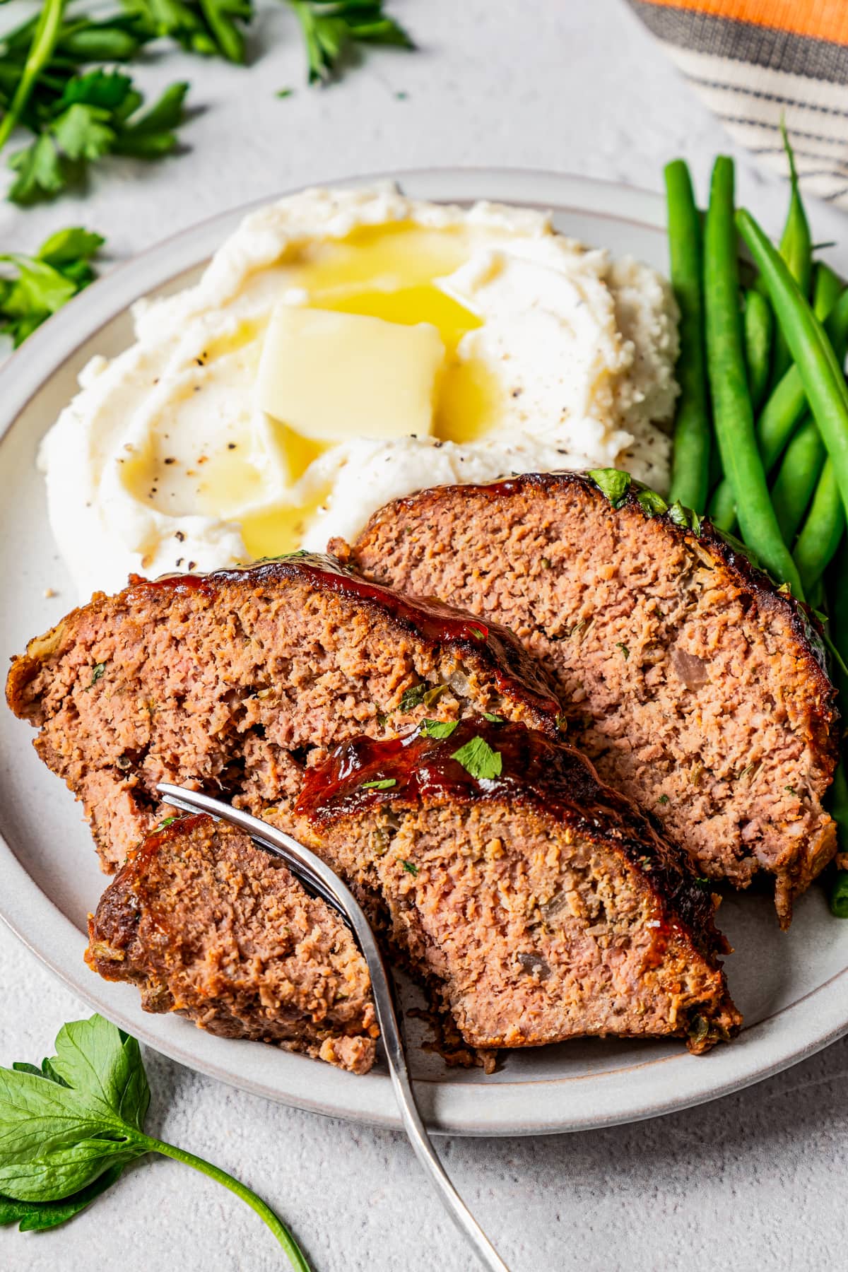 A fork taking a bite out of a slice of air fryer meatloaf on a plate with mashed potatoes and green beans.