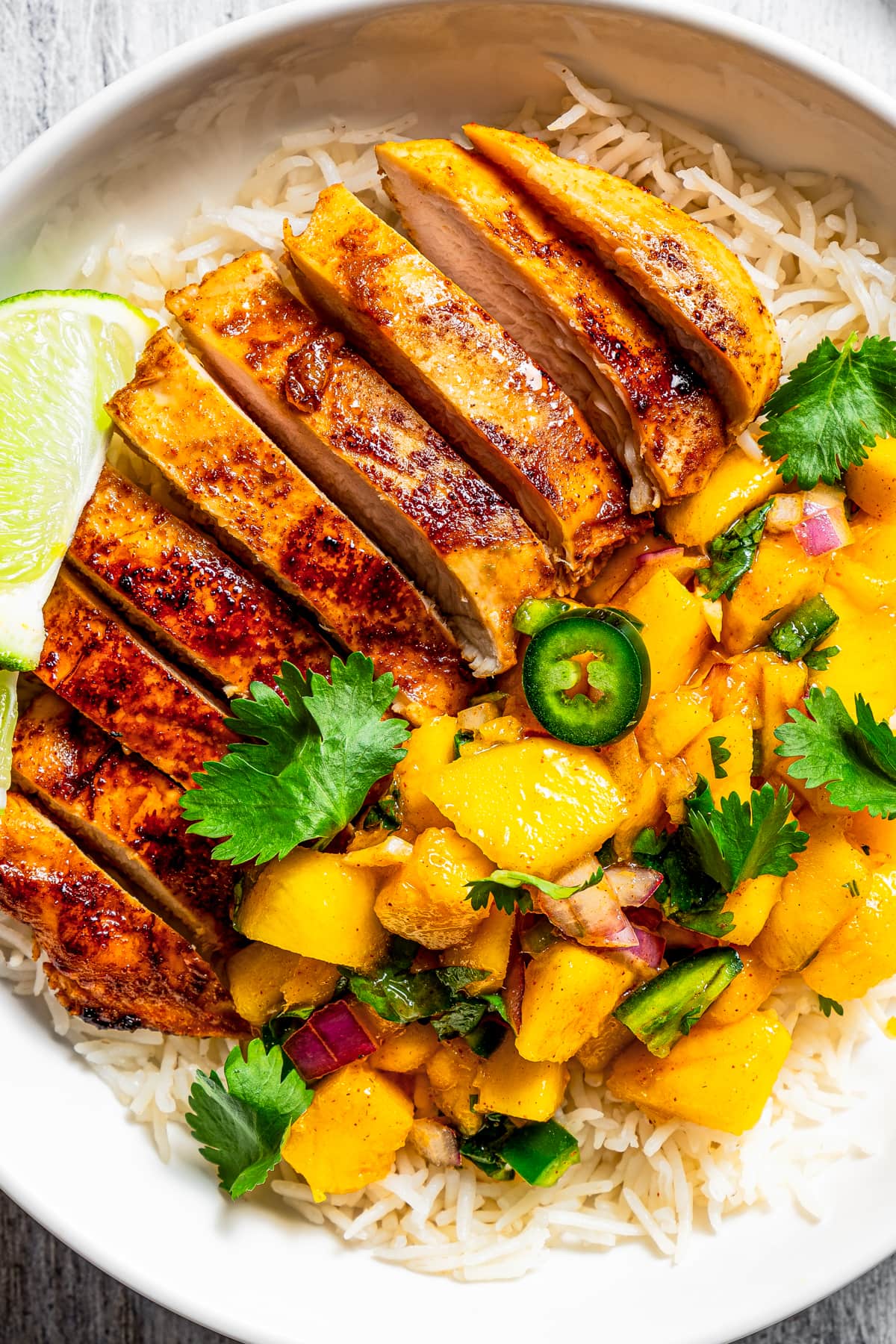 Close-up image of sliced chicken served in a bowl over rice and a side of mangoes.